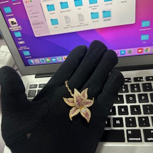 Puede incluir: Un collar con colgante floral dorado con detalles de piedras preciosas rojas y transparentes. El diseño floral presenta pétalos detallados y una cadena delicada. El collar se muestra sobre una superficie negra.