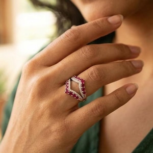 Puede incluir: Un anillo plateado con un diseño de doble banda. La banda superior presenta una serie de pequeñas piedras preciosas rojas redondas dispuestas en forma de diamante, con una fila de pequeñas piedras preciosas transparentes a cada lado. El anillo se lleva en un dedo.