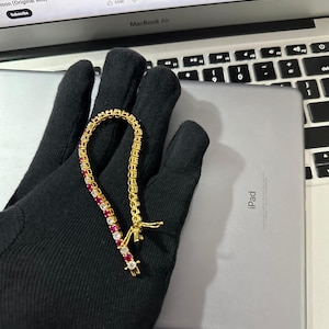 May include: A gold bracelet set with alternating red and clear gemstones. The bracelet is shown on a black surface, with a silver iPad and a MacBook Air laptop in the background.