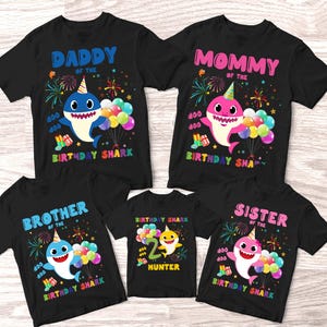 May include: Black t-shirts with colorful shark designs for a family. The shirts read "Daddy," "Mommy," "Brother," "Sister," and "Hunter" with birthday shark graphics, balloons, and fireworks. The "Hunter" shirt shows the number "2."