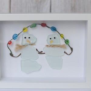Pode incluir: Uma peça de arte de parede emoldurada com dois bonecos de neve feitos de vidro do mar branco. Cada boneco tem um cachecol de fibra bege, olhos de ponto preto e um nariz laranja. Eles seguram um fio de contas coloridas e translúcidas acima.