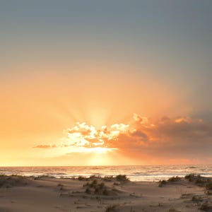 May include: A coastal landscape featuring sand dunes with sparse vegetation in the foreground, leading to an ocean horizon under a bright sunset sky. The sun is partially obscured by clouds, casting golden light and rays across the atmosphere.