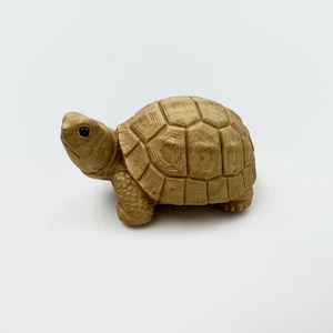 May include: A small, tan-colored turtle figurine with a detailed shell pattern. The turtle has a textured appearance and a small, black eye. The figurine is made of a solid material and sits on a white surface.