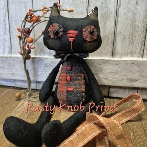 May include: A black fabric cat doll with a textured orange and grey plaid belly, featuring stitched facial details, dark button eyes, and a ruffled neck collar. The doll has long limbs and is accompanied by a length of orange ribbon.