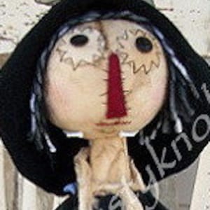 May include: A handcrafted scarecrow doll with a round, stitched face, button eyes, and a red felt nose. It wears a black hat with a sheer bow and a black jacket over a plaid shirt. The doll has straw hair and a stitched mouth.