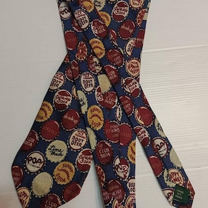 以下が含まれることがあります： 紺色のネクタイ。赤、黄、白のヴィンテージソーダボトルのキャップが繰り返されるパターン。手作りのネクタイで、様々なソーダブランド名が特徴です。