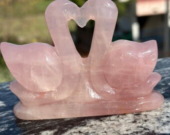 May include: A pink rose quartz sculpture of two swans forming a heart shape. The swans are carved from a translucent, light pink stone and rest on a rounded base. The sculpture is displayed outdoors in natural light.
