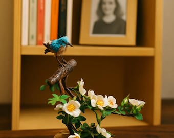 May include: A decorative sculpture of a blue and brown bird perched on a branch with white flowers and green leaves. The sculpture is on a round, cork base. A wooden bookshelf and framed photo are in the background.