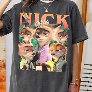 May include: Dark gray t-shirt featuring a graphic of Nick Wilde from Zootopia. The design includes the name "Nick Wilde" in orange and the character's image. The shirt is a casual, oversized fit.