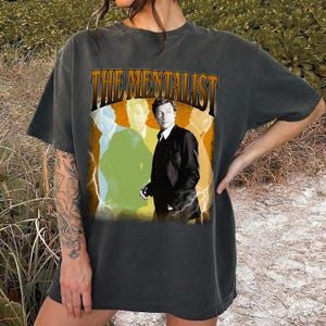 May include: Dark gray t-shirt featuring the text "THE MENTALIST" in an orange and brown font. The graphic includes a man in a black suit with a blurred image of the same man in the background, with yellow and green accents and lightning bolts.