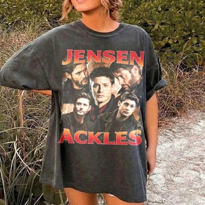 May include: Dark gray t-shirt featuring a group of men with the text "JENSEN ACKLES" in a gradient orange and red font. The shirt has a vintage, worn look.