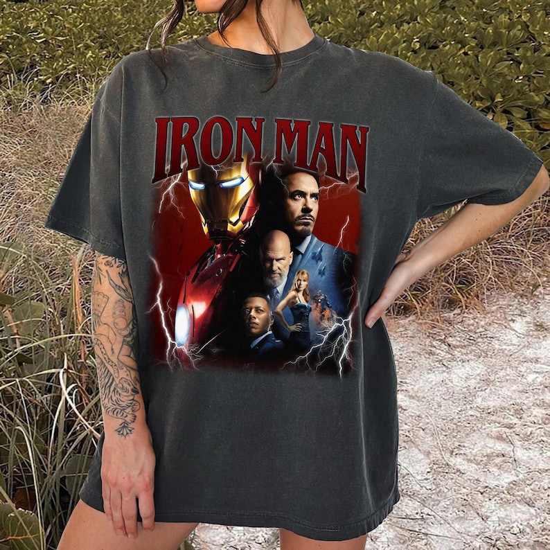 May include: Dark gray t-shirt featuring the text "IRON MAN" in red, with a graphic of the Iron Man character and other characters from the movie. The graphic is set against a red background with lightning bolts.