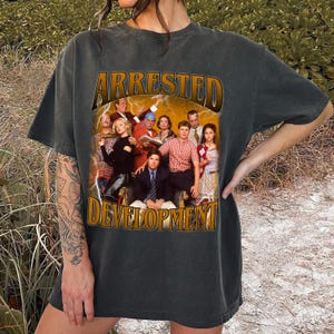Puede incluir: Camiseta gris oscuro con el texto "ARRESTED DEVELOPMENT" en fuente dorada desgastada. La camiseta muestra una foto grupal del elenco con un diseño de rayo. La camiseta es de estilo vintage.