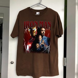 May include: Brown t-shirt featuring the text "IRON MAN" in red, with a graphic of Iron Man's helmet and several characters. The design includes lightning bolts and a red background.