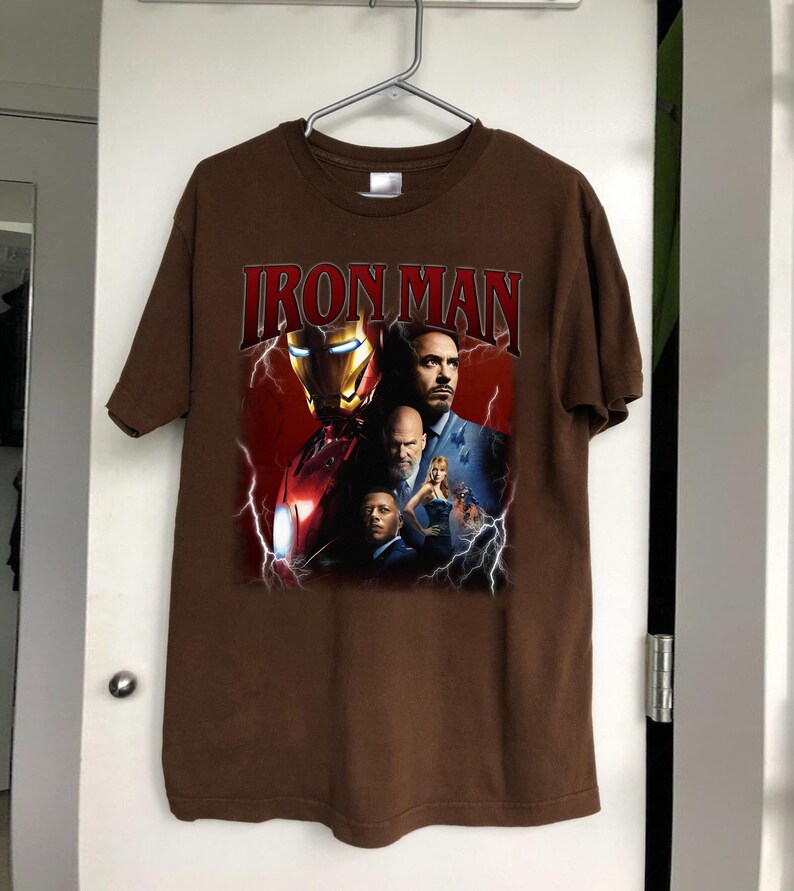 May include: Brown t-shirt featuring the text "IRON MAN" in red, with a graphic of Iron Man's helmet and several characters. The design includes lightning bolts and a red background.