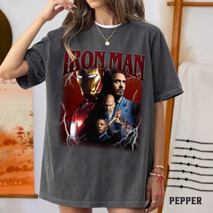 May include: Dark gray t-shirt featuring a graphic of Iron Man with the text "IRON MAN" in red. The graphic includes images of characters from the movie. The shirt is a casual wear item.