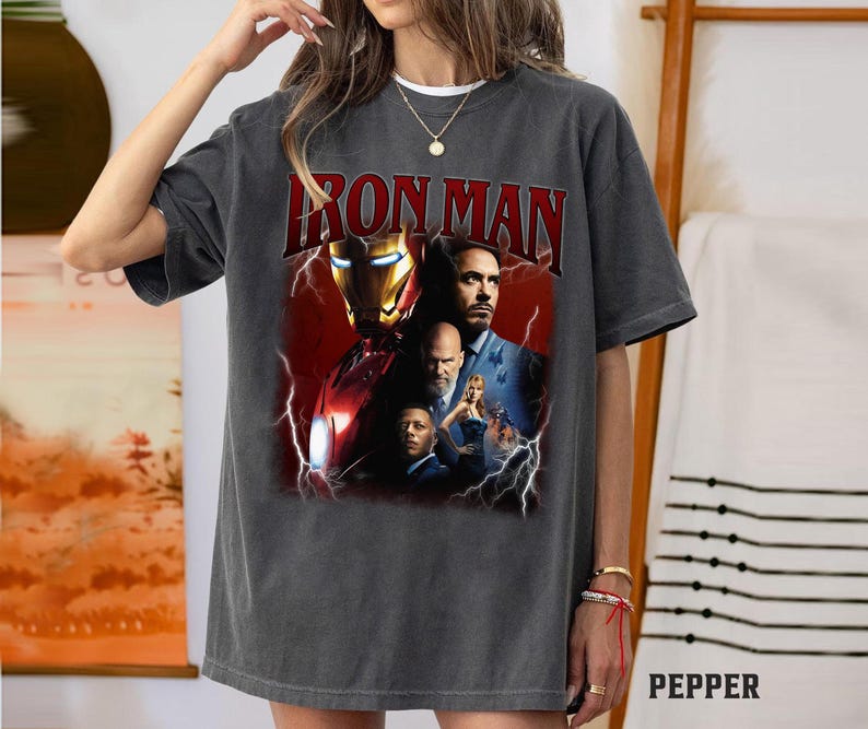 May include: Dark gray t-shirt featuring a graphic of Iron Man with the text "IRON MAN" in red. The graphic includes images of characters from the movie. The shirt is a casual wear item.