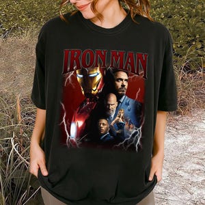 May include: Black t-shirt featuring the text "IRON MAN" in a red, distressed font above a collage of images. The collage includes the Iron Man helmet and other characters with lightning bolt graphics.