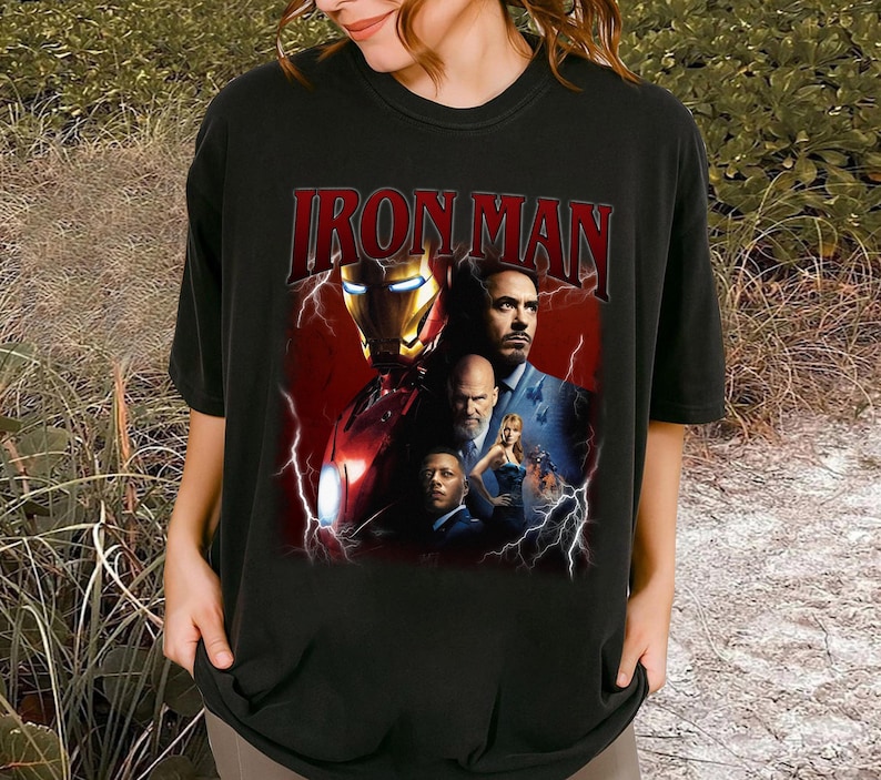 May include: Black t-shirt featuring the text "IRON MAN" in a red, distressed font above a collage of images. The collage includes the Iron Man helmet and other characters with lightning bolt graphics.