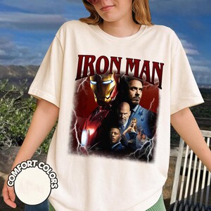 May include: Cream-colored t-shirt featuring the text "IRON MAN" in a bold, maroon font. The graphic showcases a collage of images from the movie, including Iron Man's helmet and character portraits. The shirt is labeled "COMFORT COLORS".