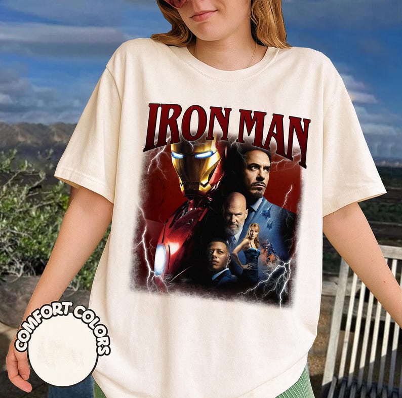May include: Cream-colored t-shirt featuring the text "IRON MAN" in a bold, maroon font. The graphic showcases a collage of images from the movie, including Iron Man's helmet and character portraits. The shirt is labeled "COMFORT COLORS".