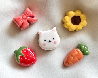 May include: A set of five small ceramic charms featuring a pink bow, a yellow sunflower with a brown center, a white cat face with pink cheeks, a red strawberry with green leaves, and an orange carrot with green leafy tops.