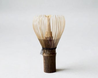 May include: A bamboo matcha whisk featuring a dark brown cylindrical handle and numerous thin, light-colored bristles arranged in a rounded, fan-like shape. A black thread is wrapped around the base of the bristles to secure them to the handle.