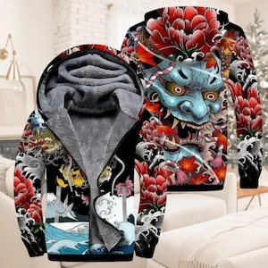 Könnte beinhalten: Schwarzer Kapuzenpullover mit Reißverschluss und japanisch inspiriertem Design. Das Design zeigt ein blaues Dämongesicht, rote Blumen und einen Drachen. Die Innenseite ist mit grauem Fleece gefüttert. Der Hoodie wird auf einem weißen Sofa präsentiert.