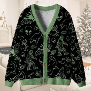 Könnte beinhalten: Schwarze Strickjacke mit grünen Zierleisten und Knopfverschluss. Der Pullover zeigt ein sich wiederholendes Muster aus grünen Illustrationen von Außerirdischen, Bigfoot, UFOs und anderen Kryptiden. Die Strickjacke wird auf einem weißen Sofa präsentiert.