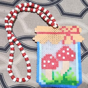 May include: A handmade necklace featuring a pendant with a pixelated mushroom design. The pendant is framed in blue and has a red and white beaded chain. The mushrooms have red caps with white spots, and the background is a light color.