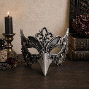 May include: A decorative silver and black raven mask with intricate details, including a bird-like beak and ornate embellishments. The mask is displayed on a dark wooden surface, with a lit candle and antique books in the background.