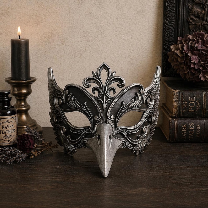 May include: A decorative silver and black raven mask with intricate details, including a bird-like beak and ornate embellishments. The mask is displayed on a dark wooden surface, with a lit candle and antique books in the background.