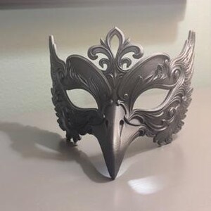 May include: A silver-colored masquerade mask with an ornate design. The mask features a bird-like beak and decorative flourishes around the eyes and forehead. The mask is made of a solid material and is designed to be worn.