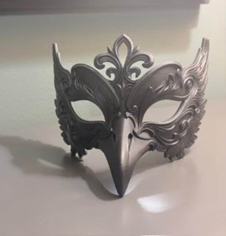 May include: A silver-colored masquerade mask with an ornate design. The mask features a bird-like beak and decorative flourishes around the eyes and forehead. The mask is made of a solid material and is designed to be worn.
