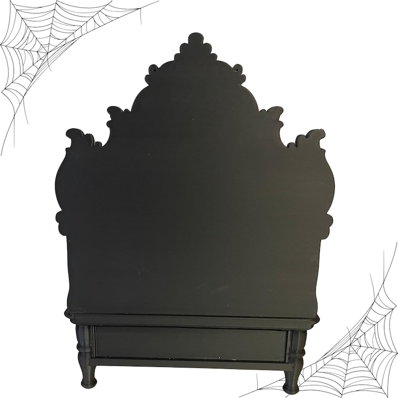 May include: A black, ornate, decorative cabinet with a drawer and decorative legs. The cabinet has a detailed, curved top. Spiderwebs are in the background. This cabinet is perfect for Halloween decor.
