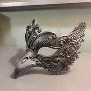 May include: Silver-colored masquerade mask with an avian design. The mask features a bird-like beak and ornate, feathered detailing around the eyes and forehead. The mask is made of a solid material and is designed to be worn for costume events.