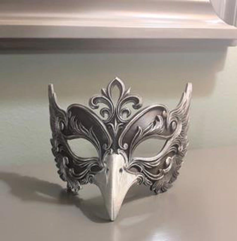 May include: A silver and white masquerade mask with an ornate, feathered design. The mask features a bird-like beak and intricate floral patterns, perfect for a costume or decorative display. The mask is made of a sturdy material and is designed to fit comfortably.