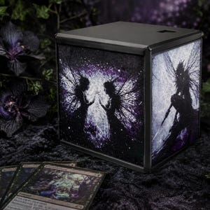 May include: A black, cube-shaped lantern with fairy artwork on the sides. The artwork features silhouettes of fairies with detailed wings and a cosmic background in shades of purple and white. The lantern is sitting on a dark, textured surface.