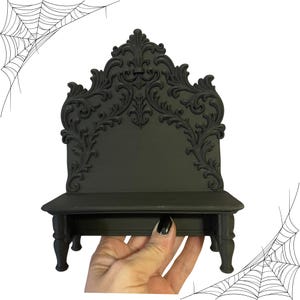 May include: A black decorative shelf with ornate scrollwork on the back and a small shelf. The shelf has a gothic or Victorian style, with a dark, matte finish. The design includes a decorative back and four small legs.