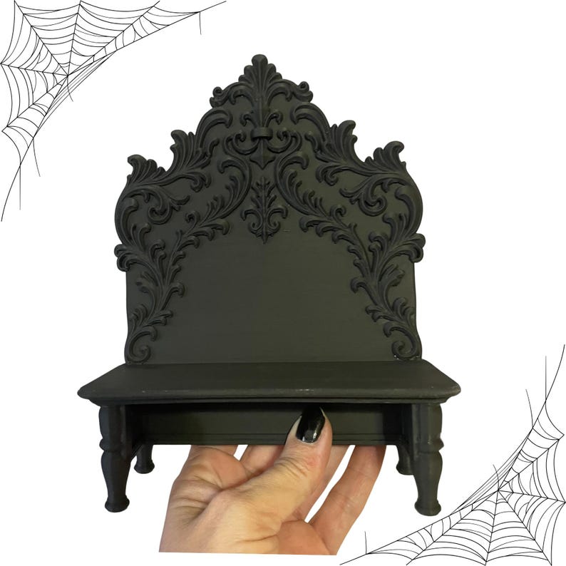 May include: A black decorative shelf with ornate scrollwork on the back and a small shelf. The shelf has a gothic or Victorian style, with a dark, matte finish. The design includes a decorative back and four small legs.