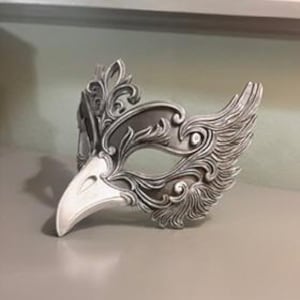May include: A silver-colored masquerade mask with a bird-like beak and ornate, swirling feather details. The mask has a detailed, textured surface and is designed to cover the upper face.