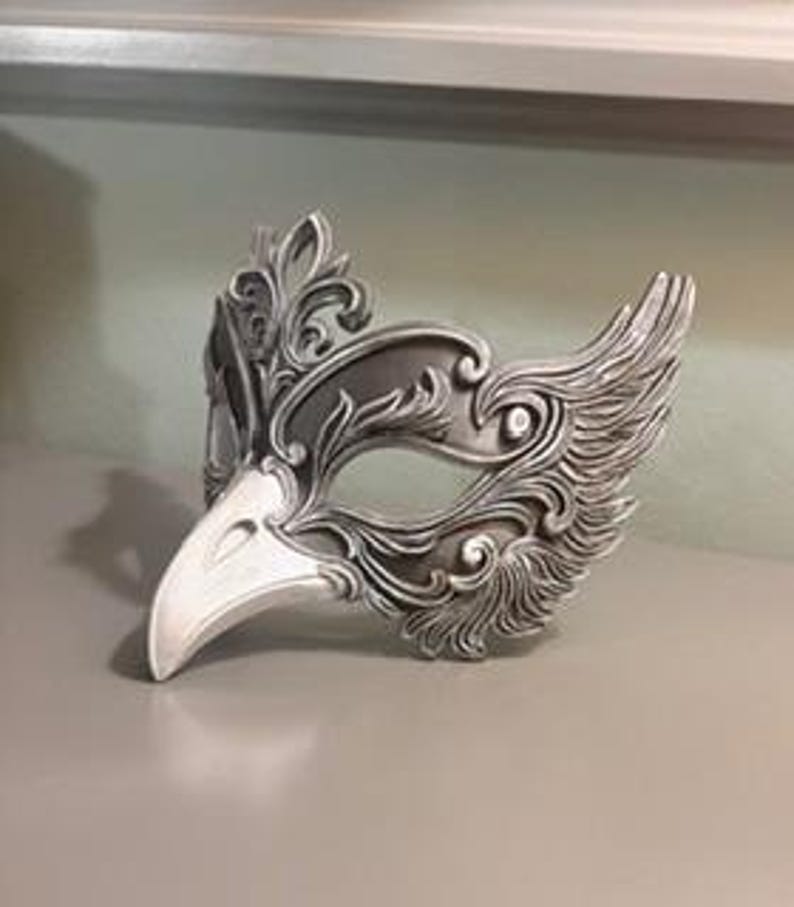 May include: A silver-colored masquerade mask with a bird-like beak and ornate, swirling feather details. The mask has a detailed, textured surface and is designed to cover the upper face.
