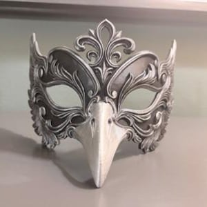 May include: A silver and white masquerade mask with an ornate, floral design. The mask features a bird-like beak and is made of a hard material. The mask is designed to cover the eyes and upper face.