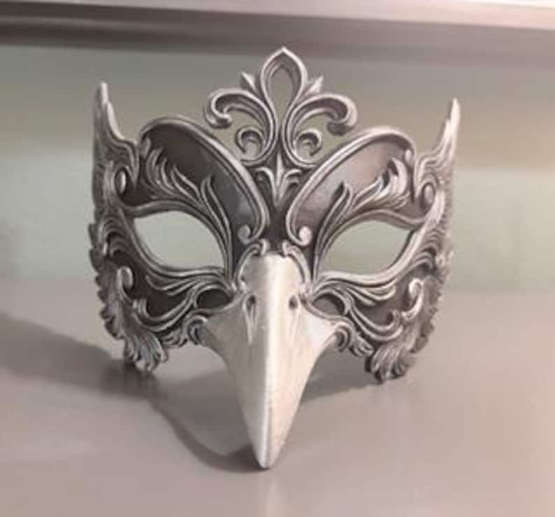 May include: A silver and white masquerade mask with an ornate, floral design. The mask features a bird-like beak and is made of a hard material. The mask is designed to cover the eyes and upper face.