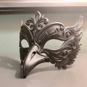 May include: A silver-colored masquerade mask with an avian-inspired design. The mask features a bird-like beak and ornate, feathered detailing around the eye and forehead areas. The mask is made of a solid material and is designed to be worn.