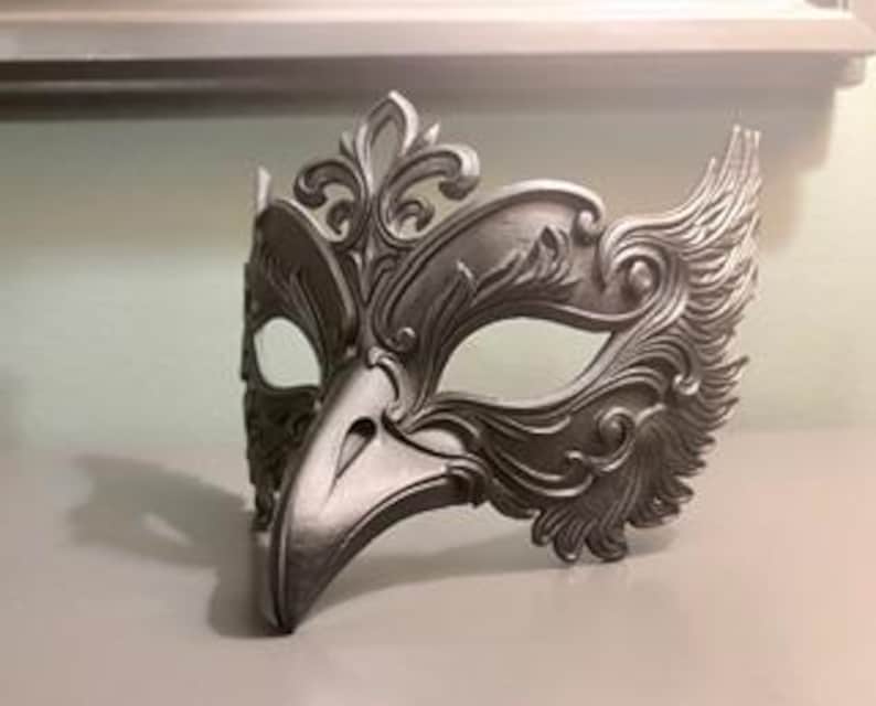 May include: A silver-colored masquerade mask with an avian-inspired design. The mask features a bird-like beak and ornate, feathered detailing around the eye and forehead areas. The mask is made of a solid material and is designed to be worn.