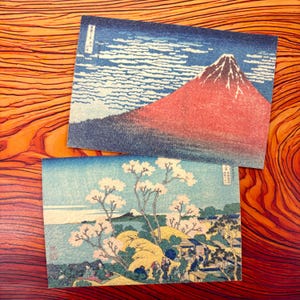 以下が含まれることがあります： 日本の木版画が描かれた2枚の長方形のポストカード。上の絵は、白い雲が浮かぶ濃い青色の空を背景にした赤い富士山を描いています。下の絵は、桜の木、人々、そして遠くの山々がある風景を描いています。