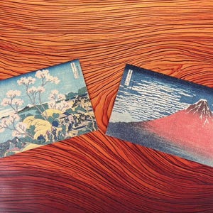 以下が含まれることがあります： 木目調の表面に2つの日本のスタイルのアートプリント。1つのプリントは桜のある風景、もう1つは曇り空の下の山を描いています。プリントは青、白、赤の色使いです。