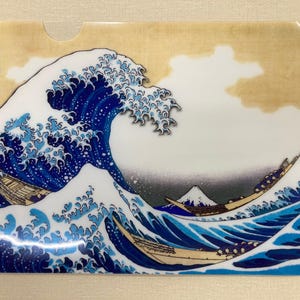 以下が含まれることがあります： 日本の象徴的な芸術作品「神奈川沖浪裏」をあしらった透明なプラスチックスリーブ。デザインは、青と白の色合いで大きな波が渦巻く様子、背景にはボートと富士山が描かれています。