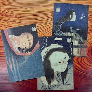 以下が含まれることがあります： 日本の木版画風のイラストが描かれた3枚の長方形のポストカード。骸骨のような生き物、長い髪の浮遊する頭部、そして暗い青色の背景に向かって煙を吐き出す女性の姿がそれぞれ描かれています。
